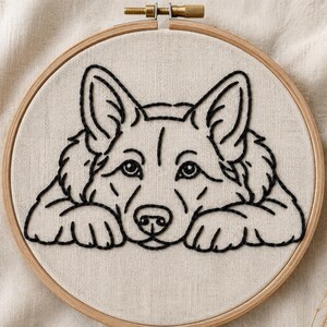 Puede incluir: Un bastidor de bordado circular de madera que contiene una tela de color blanco roto. La tela presenta un contorno de hilo negro de un perro con orejas puntiagudas y pelaje esponjoso, representado en posición de reposo con las patas delanteras.