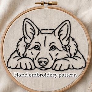 Puede incluir: Un bastidor de bordado circular de madera que contiene una tela de color blanco roto con el contorno en hilo negro de la cara y las patas de un perro. El texto "Hand embroidery pattern" está impreso en letras blancas en la parte inferior.
