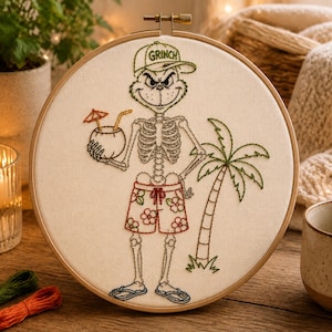 Puede incluir: Un bastidor de bordado con un esqueleto que lleva una gorra verde con el texto GRINCH, un bañador rojo con flores y chanclas. El personaje sostiene una bebida de coco con pajita y sombrilla, junto a una palmera verde bordada.