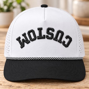 May include: A white baseball cap with a black brim and a black top button. The front features the word CUSTOM embroidered in black block letters with a curved layout. A black and white braided rope trim sits above the brim.