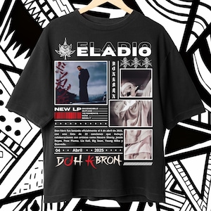 Puede incluir: Camiseta negra con el nombre "ELADIO" en blanco. La camiseta promociona un nuevo álbum, con el texto "NEW LP INVENCIBLE" y "DOH KBRON". El diseño incluye imágenes y texto, sobre un fondo con patrones en blanco y negro. La camiseta está en una percha de madera.