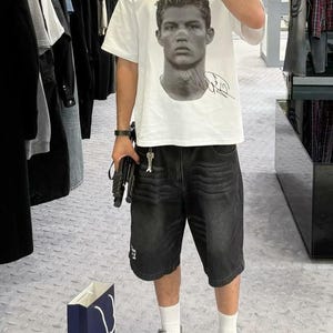 May include: A white t-shirt with a black and white image of a man's face and a signature. The man is wearing a short haircut. The person wearing the shirt is also wearing black denim shorts and white sneakers.