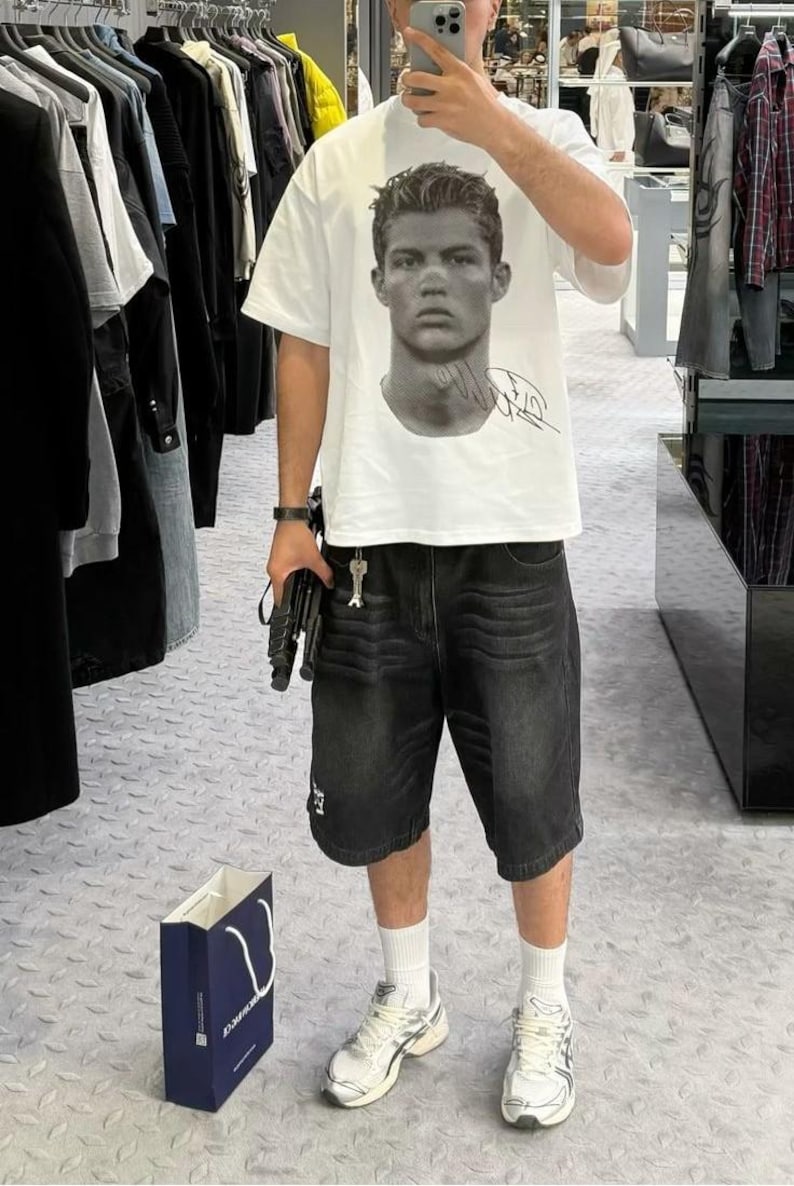 May include: A white t-shirt with a black and white image of a man's face and a signature. The man is wearing a short haircut. The person wearing the shirt is also wearing black denim shorts and white sneakers.