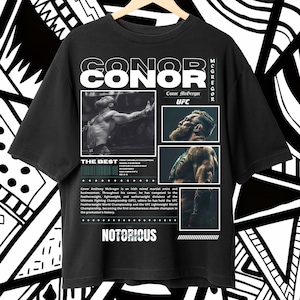 Puede incluir: Camiseta negra con el nombre "CONOR" e imágenes de Conor McGregor. La camiseta incluye el texto "UFC", "THE BEST" y "NOTORIOUS", junto con información biográfica sobre el atleta. La camiseta está colgada en una percha de madera.