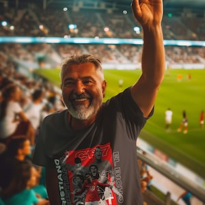 May include: A man wearing a gray t-shirt with a red and white graphic of Cristiano Ronaldo. The graphic includes the text "El Bicho" and "CR7". The man is smiling and raising his right hand in the air. He is in a stadium with a crowd of people in the background.
