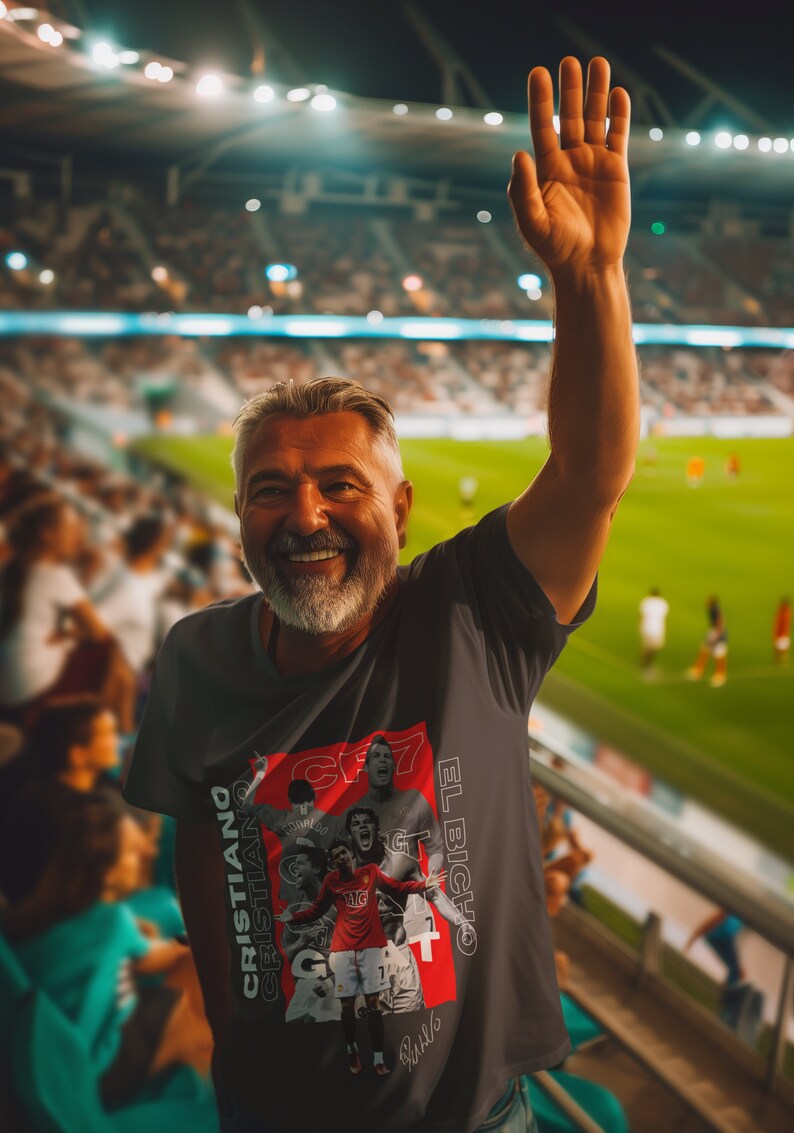 May include: A man wearing a gray t-shirt with a red and white graphic of Cristiano Ronaldo. The graphic includes the text "El Bicho" and "CR7". The man is smiling and raising his right hand in the air. He is in a stadium with a crowd of people in the background.