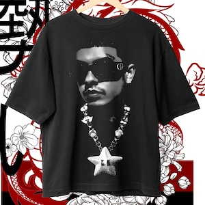 May include: Black t-shirt featuring a grayscale portrait of a person wearing sunglasses and a necklace with a star pendant. The shirt is hanging on a wooden hanger against a backdrop of floral and dragon-inspired graphics.