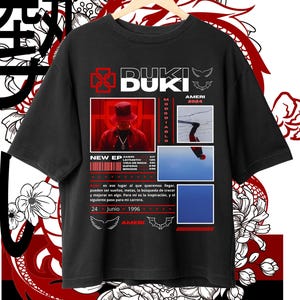 May include: Black t-shirt with a red and white graphic design featuring the text "DUKI" and "NEW EP". The graphic also includes a red bird, a red cross, and a red and white image of a person wearing a hat. The text "AMERI 2024" is also included in the graphic.