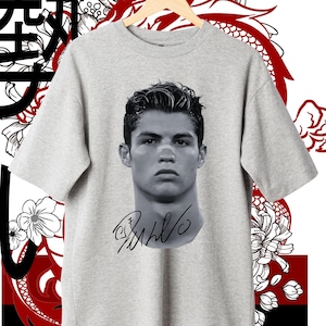 May include: A gray t-shirt with a black and white image of a soccer player's face and signature. The player's name is Cristiano Ronaldo. The t-shirt has a Japanese character on the left side of the image.