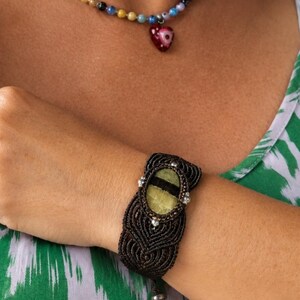 May include: A macrame bracelet featuring a central oval-shaped light green stone with a dark horizontal stripe, accented by small silver beads. A necklace with multicoloured beads and a red heart-shaped pendant with an eye design is also visible.
