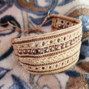 Könnte beinhalten: Ein beige-braunes Makramee-Armband mit einer Reihe von schillernden braunen Perlen.