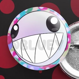 Puede incluir: Dos insignias redondas con un diseño de cara de dibujos animados. Una insignia muestra el frente con una sonrisa de dientes blancos, ojos grandes y un borde colorido. La otra muestra la parte trasera del alfiler. El fondo es negro con lunares rojos.