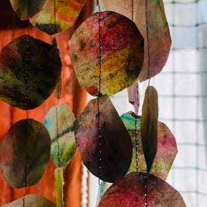 May include: A hanging mobile composed of multiple circular paper shapes in various watercolor hues including pink, yellow, green, and brown. Each paper circle is connected vertically by thin, dark brown thread stitching, creating a layered effect.