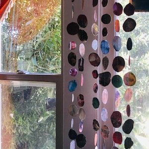 May include: A hanging mobile consisting of multiple vertical strands of thin, translucent, circular and oval-shaped slices of multicolored agate stones. The stones feature varied natural patterns and hues, including shades of blue, red, brown, and grey, suspended on thin, light-colored strings.