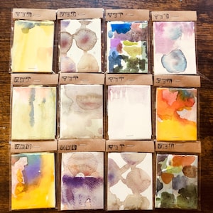 May include: Twelve rectangular watercolor paintings on paper, each individually packaged in a clear plastic sleeve with a brown paper header labeled "LEA". The paintings feature various abstract color washes, including yellow, purple, blue, orange, and earth tones, displayed in a grid arrangement.