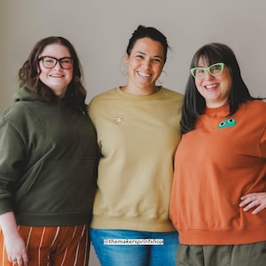 Puede incluir: Tres personas con ropa informal. Una persona lleva una sudadera con capucha verde y pantalones a rayas naranjas. Otra lleva una sudadera amarilla con un parche de zorro. La tercera lleva una sudadera naranja con un parche verde.