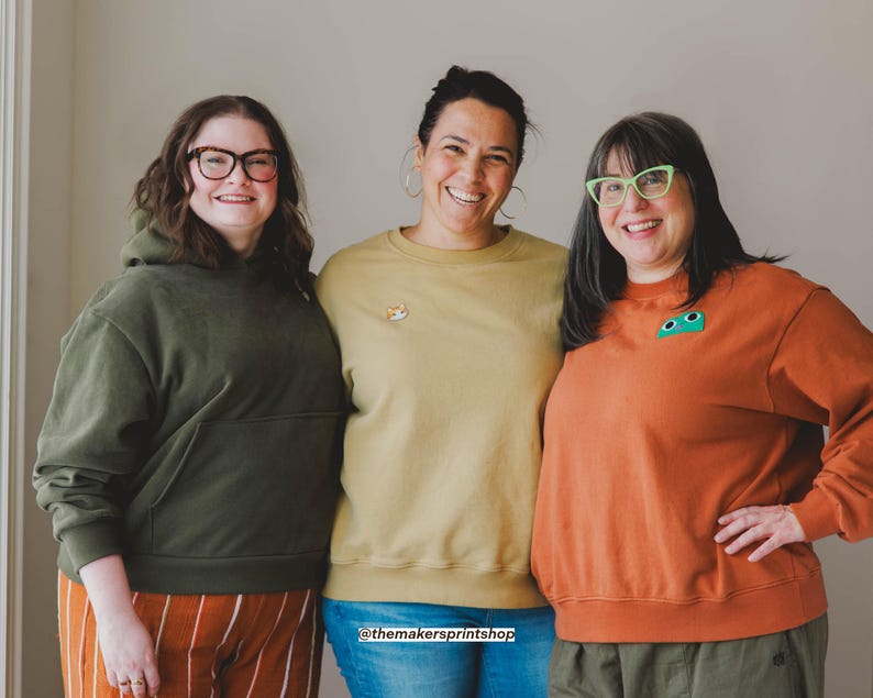 Puede incluir: Tres personas con ropa informal. Una persona lleva una sudadera con capucha verde y pantalones a rayas naranjas. Otra lleva una sudadera amarilla con un parche de zorro. La tercera lleva una sudadera naranja con un parche verde.
