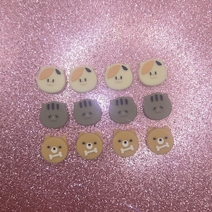May include: A collection of twelve decorative buttons with animal faces. The buttons are arranged in three rows of four. The top row features cat faces in white, tan, and black. The middle row has gray cat faces. The bottom row has tan bear faces with a bone.