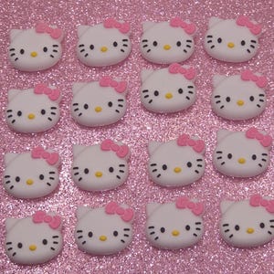May include: A collection of sixteen white Hello Kitty face-shaped embellishments with pink bows and black facial features. The embellishments are arranged on a sparkly pink surface, creating a cute and playful aesthetic.