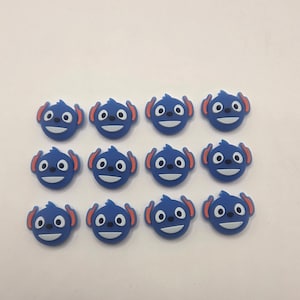 May include: Twelve blue cartoon character face charms with red ears and white facial features are arranged on a white surface. The charms have a smiling expression and are likely for crafting or jewelry making.