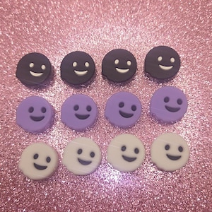 May include: An assortment of twelve smiley face charms in three colors: dark purple, light purple, and white. Each charm has a black smiling face. The charms are arranged in three rows of four.