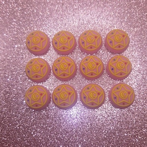 May include: A collection of twelve pink, circular resin cabochons. Each cabochon features a yellow star and crescent moon design with blue and red accents. The cabochons are arranged on a sparkly pink surface.