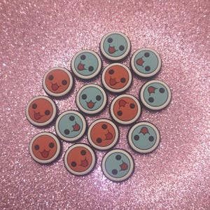 May include: A collection of round, colorful charms with smiling faces. The charms alternate between orange and light blue, each with a white border and black details. The background is a sparkly pink surface.