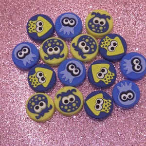 May include: A collection of round, blue and yellow decorative buttons with cartoon octopus and number eight designs. The buttons are arranged on a pink glitter surface, creating a playful and colorful display.