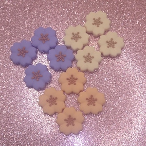 May include: An assortment of flower-shaped embellishments in shades of lavender, cream, and peach. Each flower has a contrasting center, and they are arranged on a sparkly pink surface. These decorative elements are suitable for crafts and DIY projects.