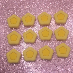 May include: Twelve yellow star-shaped embellishments with a lighter yellow star design in the center. The embellishments are arranged in three rows of four on a sparkly pink background. These are likely craft supplies.