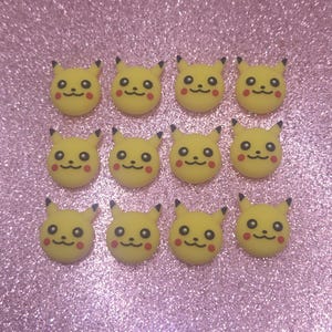 May include: A collection of twelve yellow Pikachu-shaped embellishments. Each has black-tipped ears, small black eyes, a red cheek, and a smiling mouth. The embellishments are arranged on a sparkly pink surface.