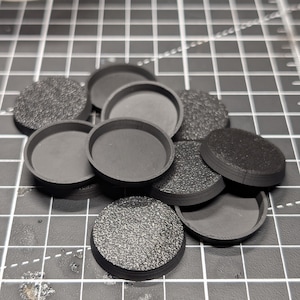 Könnte beinhalten: Eine Sammlung schwarzer, runder Kunststoffsockel, einige strukturiert und andere glatt, für Miniaturfiguren. Die Sockel sind für Tabletop-Spiele konzipiert. Die strukturierten Sockel haben eine körnige Oberfläche, während andere glatt sind.