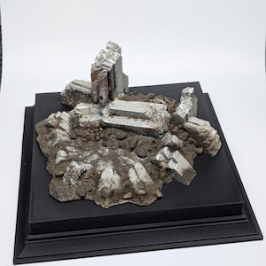 Puede incluir: Un diorama en miniatura de una estructura en ruinas, con fragmentos de piedra grises y angulares y escombros. La escena está ambientada sobre una base marrón texturizada con pequeños guijarros, todo montado sobre una plataforma negra escalonada.