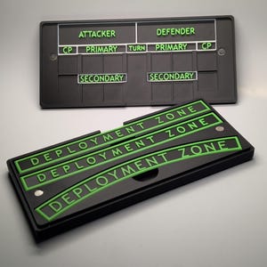 May include: Two black game boards with green lettering. The top board reads "ATTACKER" and "DEFENDER" with other labels. The bottom board has "DEPLOYMENT ZONE" repeated three times. The boards are rectangular.