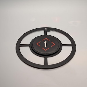 Puede incluir: Diana de metal negro con diseño circular. La diana tiene un parche central negro con una flecha roja y el número 1. La diana tiene 2,54 cm de diámetro.