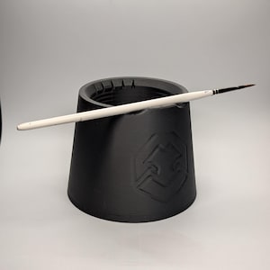 May include: A black paint brush holder with a white paint brush resting on top. The holder is cylindrical with slots around the top edge. A logo is embossed on the side of the holder.