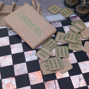Puede incluir: Un juego de mesa con piezas y una caja de color beige. La caja tiene la palabra "ORDERS" en verde. Las piezas del juego tienen bordes verdes con frases como "TAKE AIM" y "MOVE!". El juego está sobre una superficie a cuadros en blanco y negro.