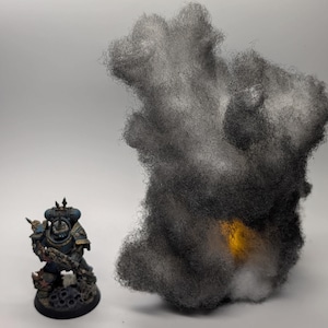 May include: A miniature figurine of a warrior in blue and gold armor, standing next to a cloud of dark gray fluff with a yellow center. The figurine is on a black base with skull details.