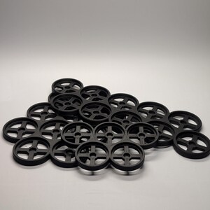 May include: A collection of black, circular objects with a cross-shaped design in the center. The objects are arranged in a pile against a white background. The objects appear to be made of plastic.