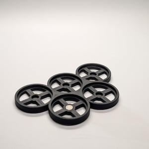 May include: A cluster of six black, circular coasters with a wheel-like design. Each coaster has a central hole and radiating spokes. A small, silver magnet is visible in the center of one coaster. The coasters are arranged on a white surface.