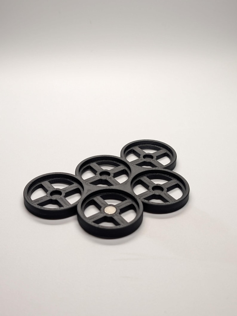 May include: A cluster of six black, circular coasters with a wheel-like design. Each coaster has a central hole and radiating spokes. A small, silver magnet is visible in the center of one coaster. The coasters are arranged on a white surface.