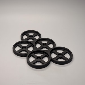 May include: A cluster of five black, circular coasters. Each coaster has a wheel-like design with a central hole and four spokes. The coasters are arranged in a slightly overlapping pattern against a plain background.