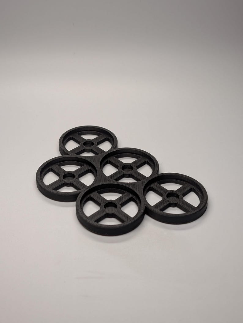 May include: A cluster of five black, circular coasters. Each coaster has a wheel-like design with a central hole and four spokes. The coasters are arranged in a slightly overlapping pattern against a plain background.