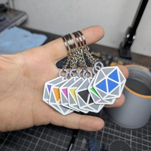 Puede incluir: Una colección de seis llaveros geométricos con forma de dados. Cada llavero presenta un diseño gris y blanco con un interior de diferente color: morado, naranja, rosa, amarillo, negro y azul. Los llaveros están sujetos a anillas plateadas.