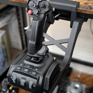 Könnte beinhalten: Schwarzer Flugsimulator-Joystick mit mehreren Knöpfen, Schaltern und einem roten Knopf. Der Joystick ist an einer schwarzen Halterung befestigt, die an einem Holzschreibtisch befestigt ist. Das Gerät hat einen strukturierten Griff und verschiedene Bedienelemente.