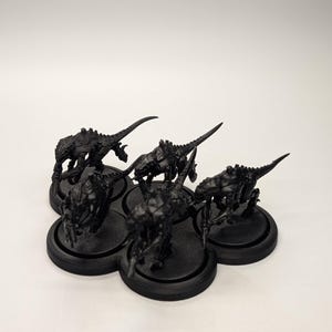 May include: A cluster of six black miniature figurines, each depicting a creature with a spiky, armored body and a long, pointed tail. The figurines are arranged on a black, multi-holed base, set against a white background.