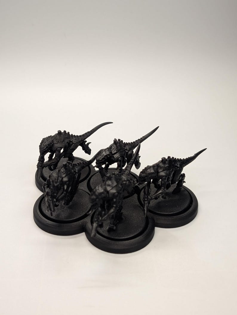 May include: A cluster of six black miniature figurines, each depicting a creature with a spiky, armored body and a long, pointed tail. The figurines are arranged on a black, multi-holed base, set against a white background.