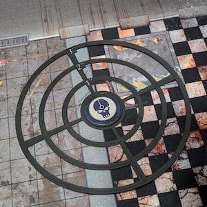 May include: A black, circular game piece with concentric rings and a central emblem featuring a skull. The piece has markings indicating measurements in inches. The background shows a checkered pattern and a textured surface.