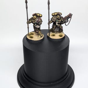 May include: Two miniature figures of armored soldiers, one holding a spear and the other a weapon, stand on a black cylindrical base. The figures are painted in gold and red tones, with detailed armor and weaponry.