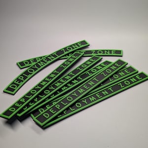 May include: A collection of black rectangular patches with the words "DEPLOYMENT ZONE" in green lettering. The patches have a green border and are arranged in a slightly curved pattern against a light background.
